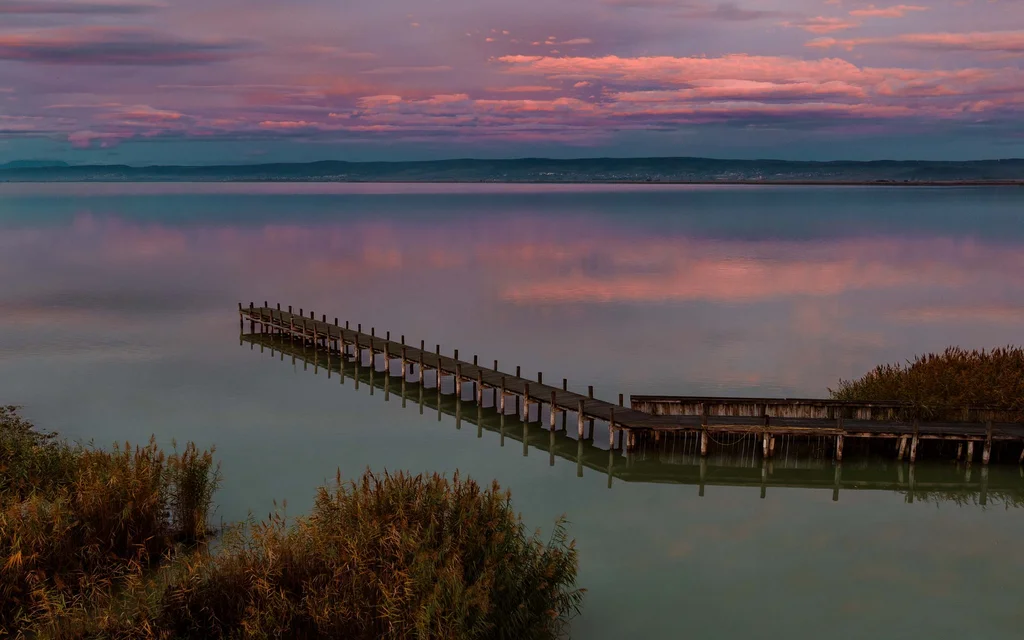 Steg auf ruhigem See bei Sonnenuntergang mit rosa bewölktem Himmel