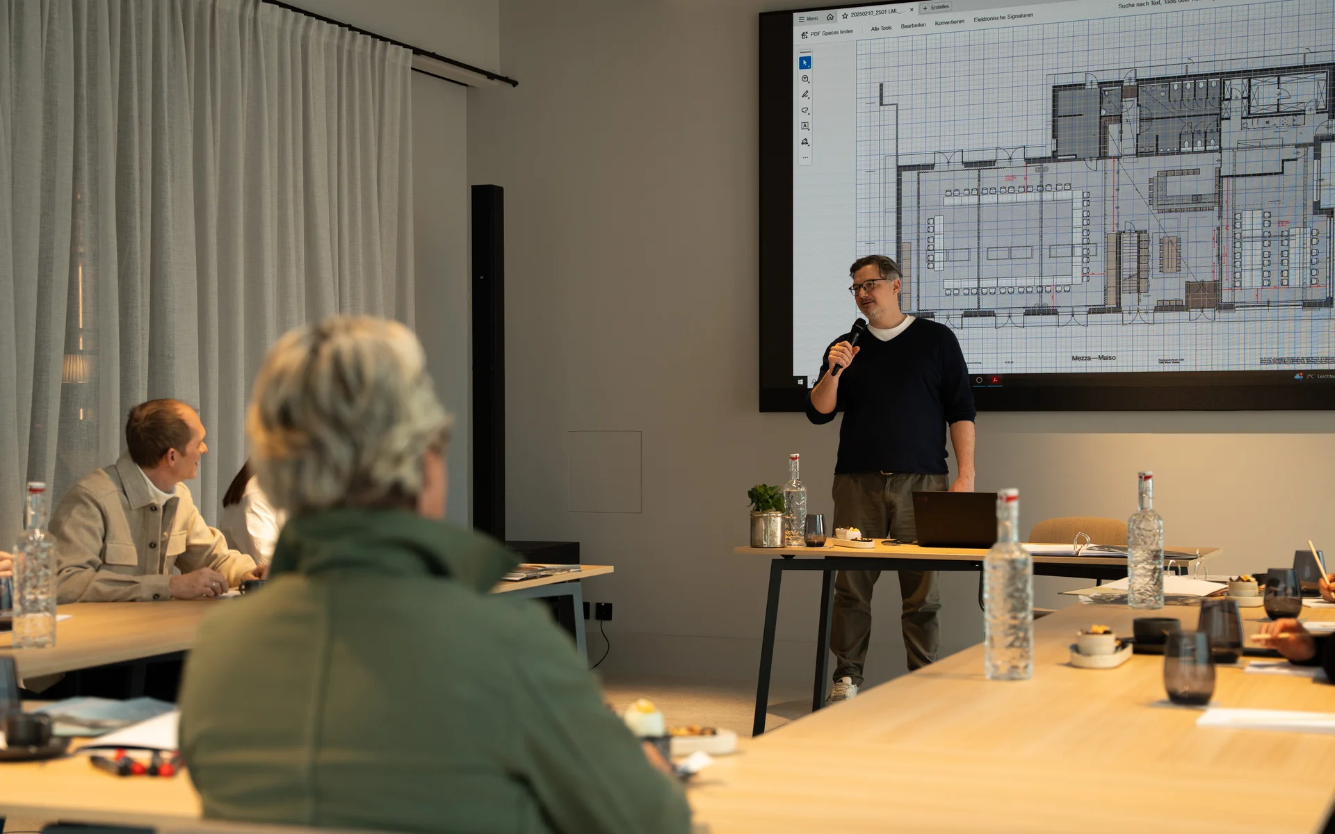 Person hält Präsentation mit Bauplan vor einem Konferenztisch mit Zuschauern