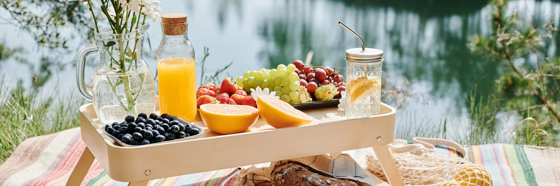 Picknick mit Obst, Brot, Getränken und Blumen auf Decke am See
