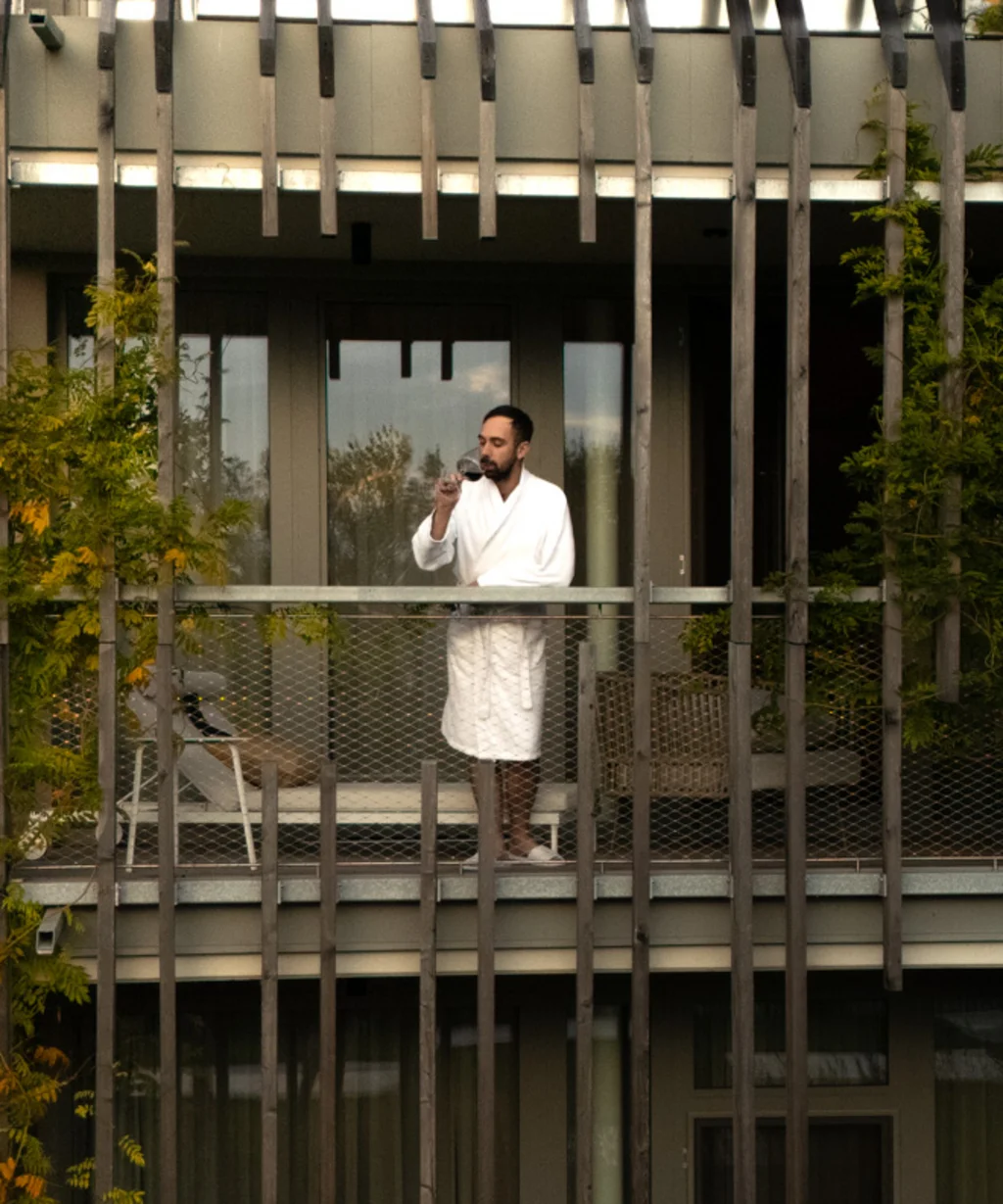 Mann im Bademantel trinkt Getränk auf Balkon eines begrünten Gebäudes