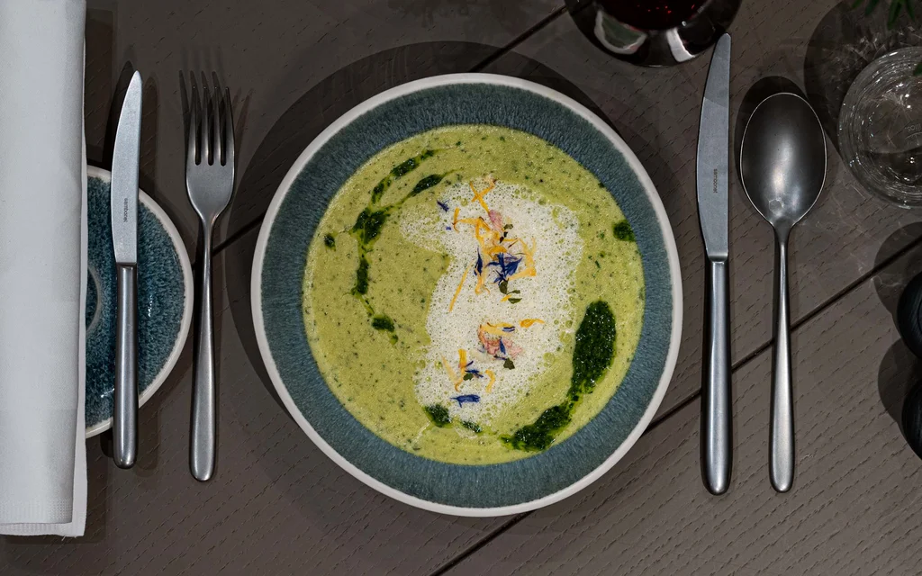 Grüne Suppe mit Schaum und Kräutern auf blauem Teller, Besteck und Glas daneben