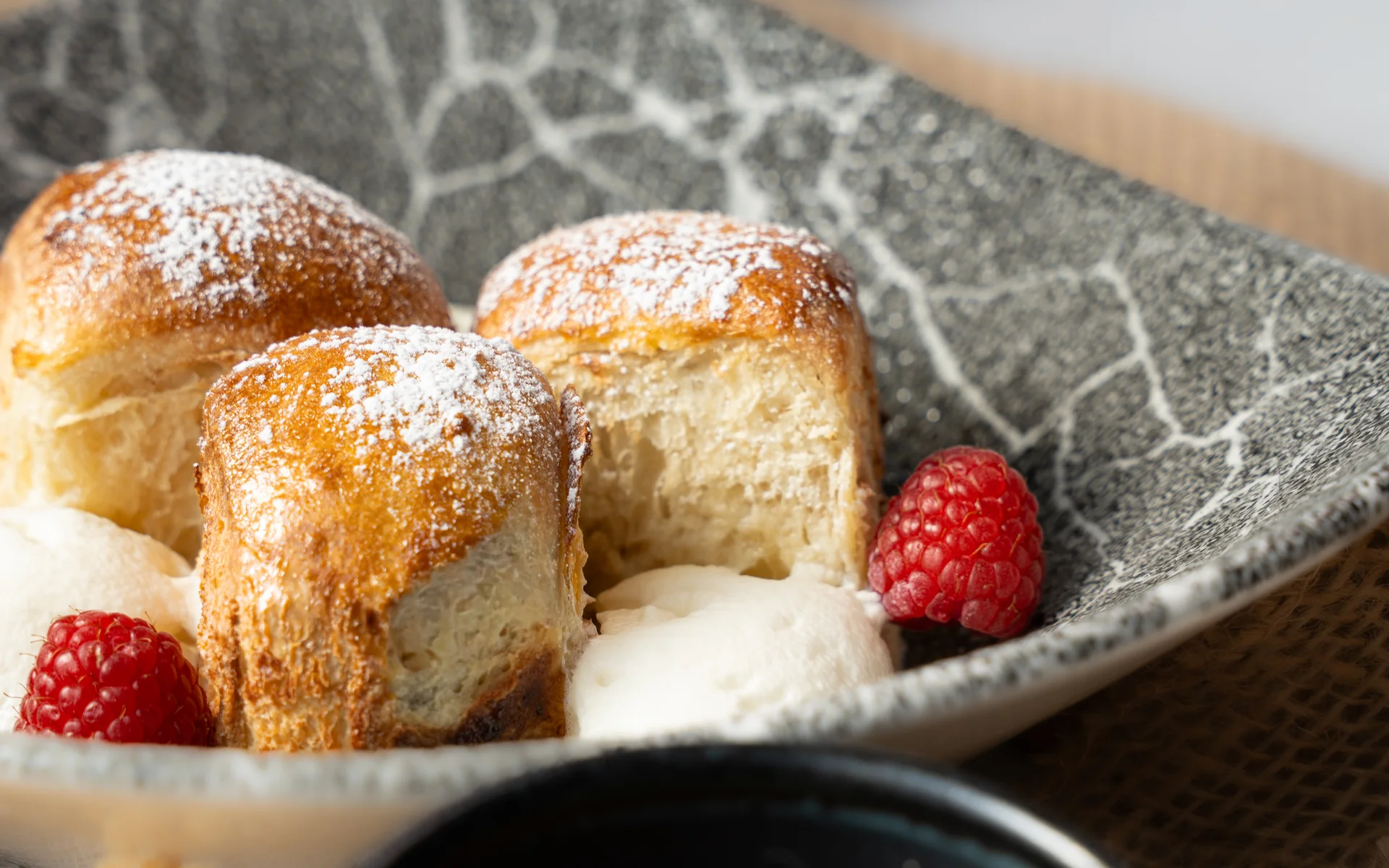 Drei leicht gezuckerte Gebäckstücke mit Sahne und Himbeeren auf einem grauen Teller